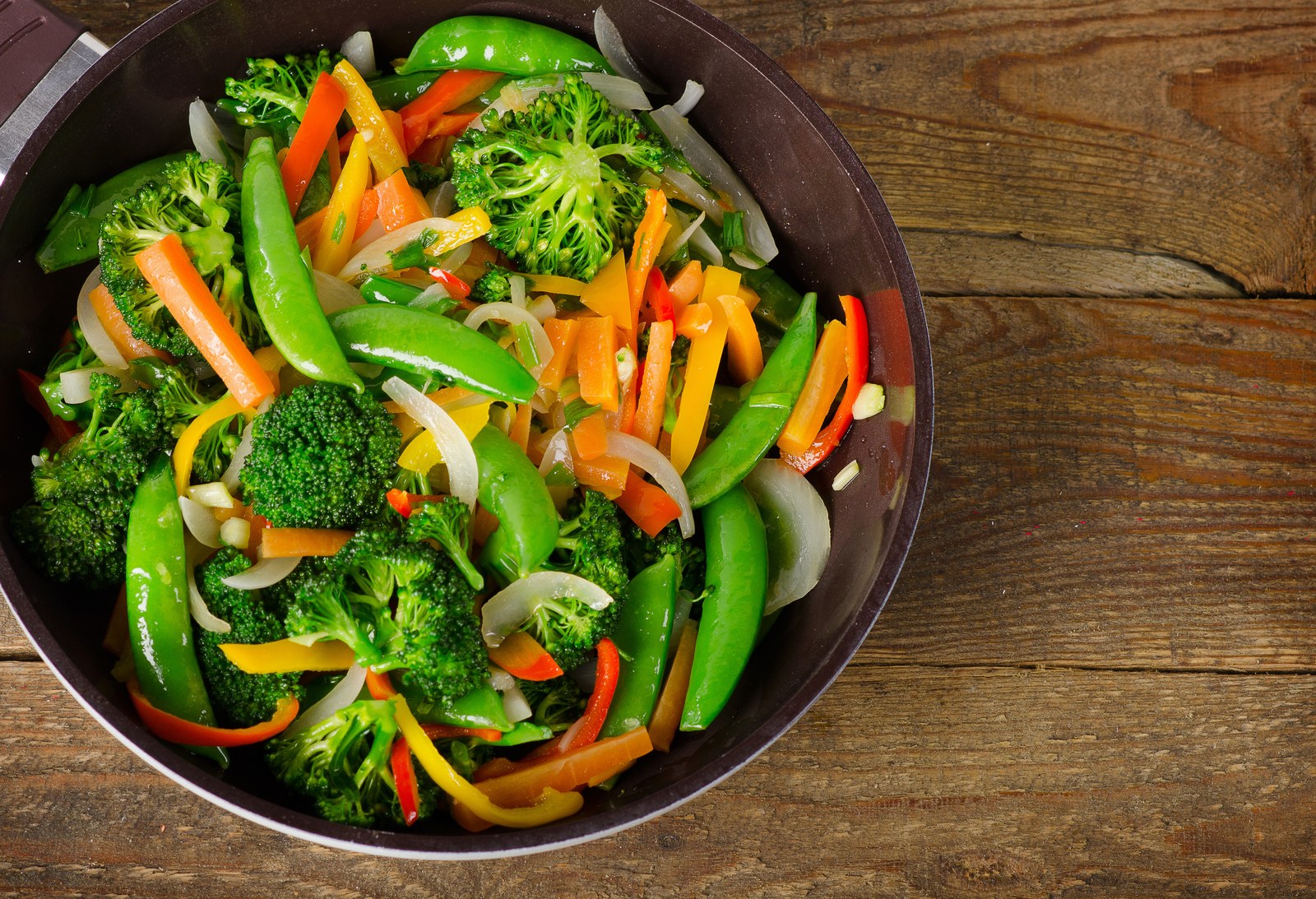 Colorful vegetable stir-fry with bright green snap peas, broccoli florets, orange carrots, yellow and red bell peppers, sliced onions in dark wok