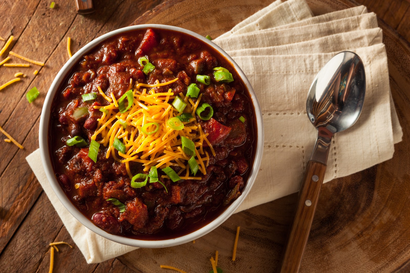 Vegetarian bean chili with dark red kidney beans, tomato sauce, topped with shredded cheddar cheese, sliced green onions in white bowl on wooden table