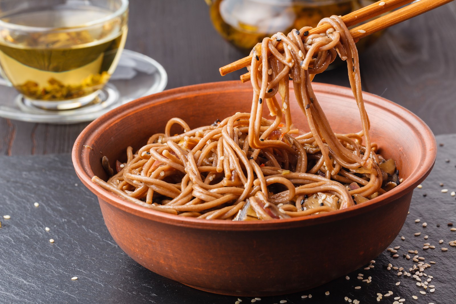 Japanese zaru soba cold buckwheat noodles held in chopsticks over terracotta bowl, sesame seeds, served with green tea on dark slate